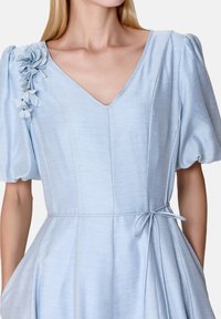 Robe bleu clair avec un décolleté en V, des manches bouffantes, un accent floral sur l'épaule gauche et un détail de taille nouée. Tissu lisse et texturé.