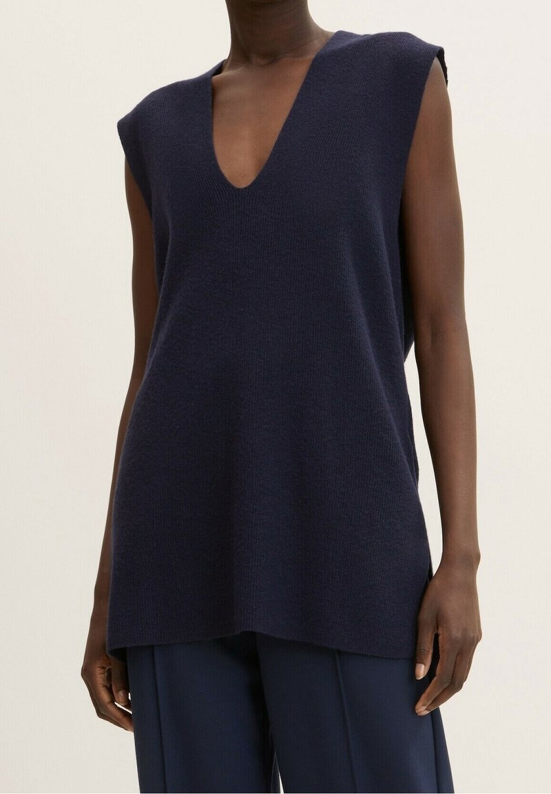 Veste en tricot sans manches bleu marine foncé avec un col en V profond portée sur un pantalon large assorti, sur une personne debout.