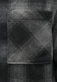 Μαύρο και γκρι πουκάμισο από φανέλα με καρό σχέδιο, διαθέτει τσέπη στο στήθος και υφή υφάσματος με αντιθετικά μοτίβα από μεγάλα και μικρά τετράγωνα.