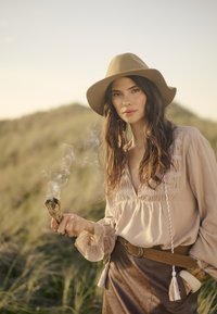 Helles beige Blusenshirt mit Rüschendetails, braut Lederrock, weitkrempiger beiger Hut, hält ein brennendes Salbeibündel. Sanfter Gras-Hintergrund.