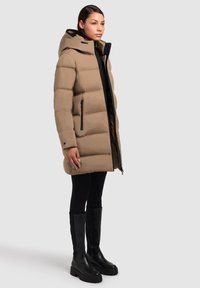 Abrigo tipo puffer en beige con secciones acolchadas horizontales, un zipper negro y capucha. Combinado con leggings negros y botas altas hasta la rodilla.