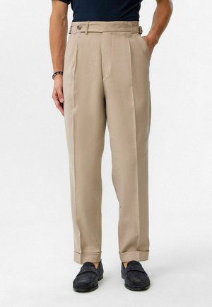 Beige maßgeschneiderte Hose mit hoher Taille, seitlichen Falten und Knopfakzenten, kombiniert mit dunklen Slippern. Glatter Stoff mit einem klaren Finish.