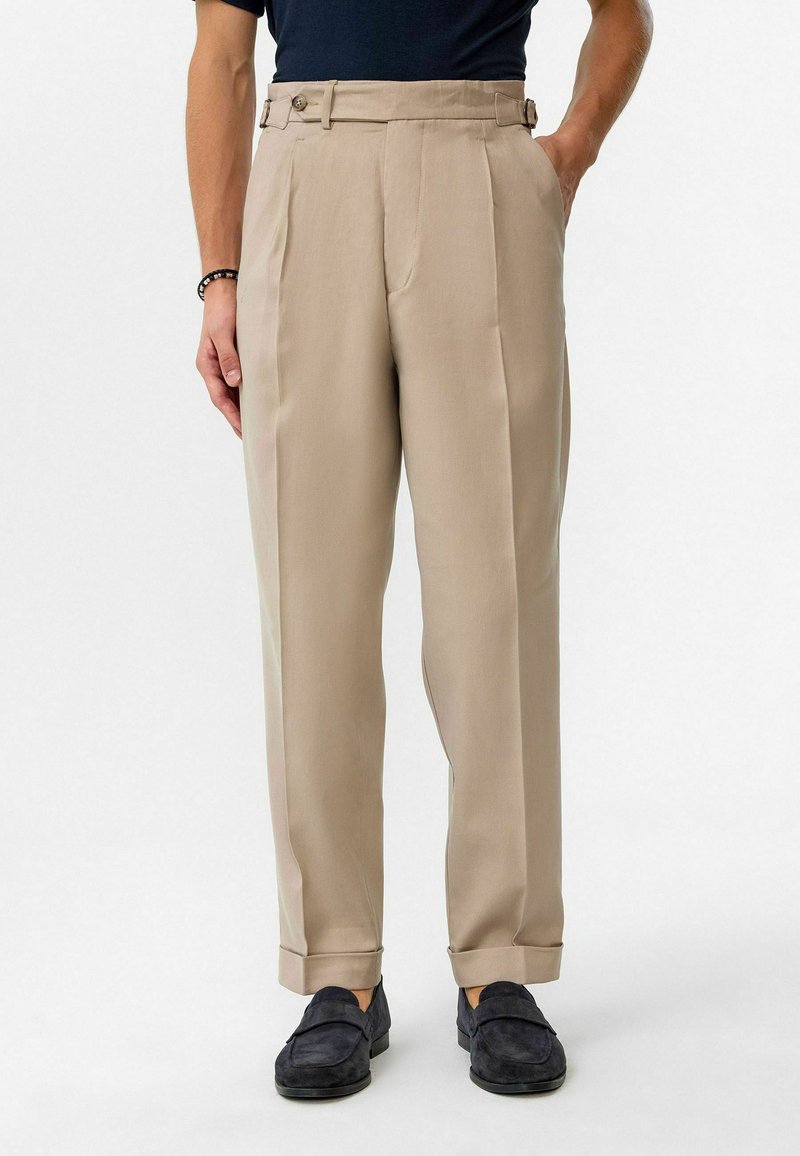 Beige op maat gemaakte broeken met een hoge taille, zijsplitten en knopenaccenten, gecombineerd met donkere loafers. Glad materiaal met een strakke afwerking.
