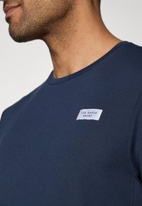 Sötétkék, rövid ujjú póló, mely texturált anyagból készült, a mellkasán található fehér címkével és a "TED BAKER SPORT" logóval.