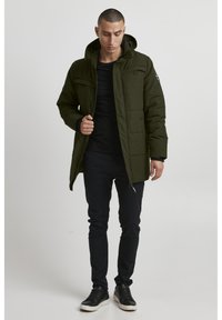 Grønn polstret jakke med glidelås og knappeåpning, hette og to brystlommer. Brukt over en svart skjorte og kombinert med svarte bukser og sneakers.