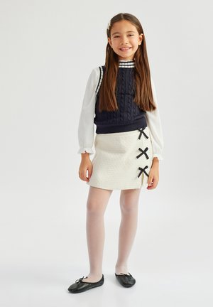 Young girl standing, wearing a navy cable knit vest over a white blouse, cream skirt with three black bows, white tights, and black flats.