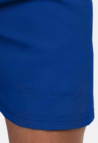 Blauwe zwembroek met een gladde textuur, subtiele glans en geborduurd "Reichstadt" logo nabij de zoom. Eenvoudig, gestroomlijnd ontwerp.