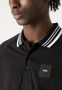 Camisa polo negra con un cuello a rayas, dos botones en el cuello y un parche negro texturizado con la palabra "BOSS" en blanco.