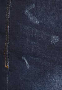 Tela de denim en azul oscuro con una superficie ligeramente texturada. Presenta costuras amarillas a lo largo de la costura y marcas suaves blancas desgastadas.