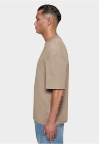 Beige cotton t-shirt, short sleeves, crew neck, relaxed fit. Worn with light blue jeans, side profile shown against a plain light background.