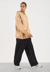 Joven caminando, vistiendo una sudadera grande color beige, pantalones negros holgados y zapatillas de correr blancas, contra un fondo claro y liso.