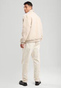 Beige fleece jacket with a high collar and ribbed cuffs, paired with off-white trousers and black shoes, shown from the back.
