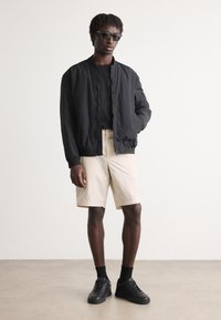 Svart bomberjacka med texturerat tyg, svart t-shirt, beige shorts och svarta sneakers. Modell som bär mörka solglasögon. Minimalistisk bakgrund.