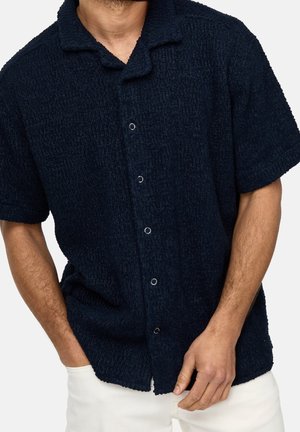 Homme portant une chemise bleu marine texturée à manches courtes avec col et boutons, et un pantalon blanc, main dans la poche, sur un fond uni.