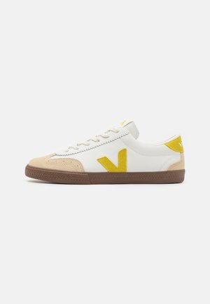 Sneaker bianco con accenti gialli e beige, caratterizzato da una punta in suede testurizzato, design con lacci e suola in gomma con un battistrada dal motivo.