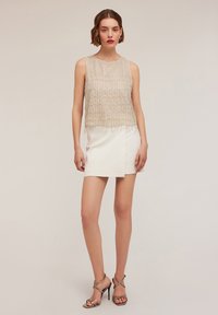 Kralen mouwloze top in roomkleur, met een textuurontwerp en franjes aan de randen, gecombineerd met een strakke witte minirok en metallic hakken met bandjes.
