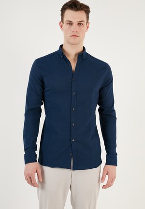 Camisa de botones azul marino con mangas largas, un cuello estructurado y una tela texturizada. Presenta botones negros y un corte entallado.