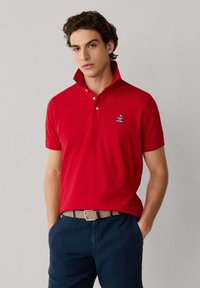 Jeune homme portant un polo rouge au col relevé, un pantalon bleu marine et une ceinture tissée beige, debout les mains dans les poches.