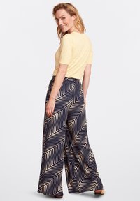 Geel shirt met korte mouwen en textuurelementen, gecombineerd met marineblauwe wijde broek met een golvend crèmekleurig patroon. Het model staat zijwaarts, met een glimlach.