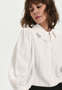 Weiße, strukturierte Bluse mit einem erhabenen Streifenmuster, einem großen Kragen und Puffärmeln, mit einem Knopfverschluss am Hals.
