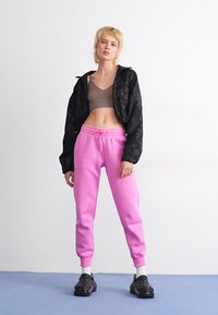 Svart mønstrete cropped jakke over en grå topp, kombinert med rosa joggebukser og svarte slip-on sko. Glatte teksturer og uformelt design.