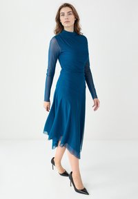 Robe midi teal avec un corsage ajusté, de longues manches transparentes et une jupe fluide dotée d'un ourlet asymétrique. Associée à des talons noirs.