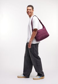Bolsa de mensajero acolchada color burdeos con una correa ajustable, forma rectangular, textura suave y un sutil logo de la marca en un lado.