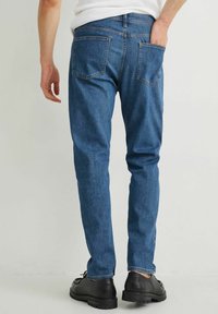 Jeans de mezclilla azul con corte recto, que presentan dos bolsillos traseros; llevados con zapatos negros, vistos desde atrás contra un fondo neutro.