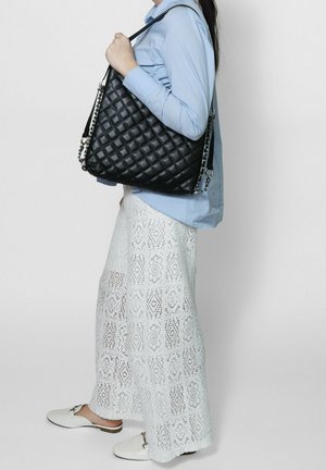 Persona con indosso una camicia blu e pantaloni bianchi a gamba larga con motivi, che tiene una grande borsa a tracolla nera trapuntata con cinghie a catena.