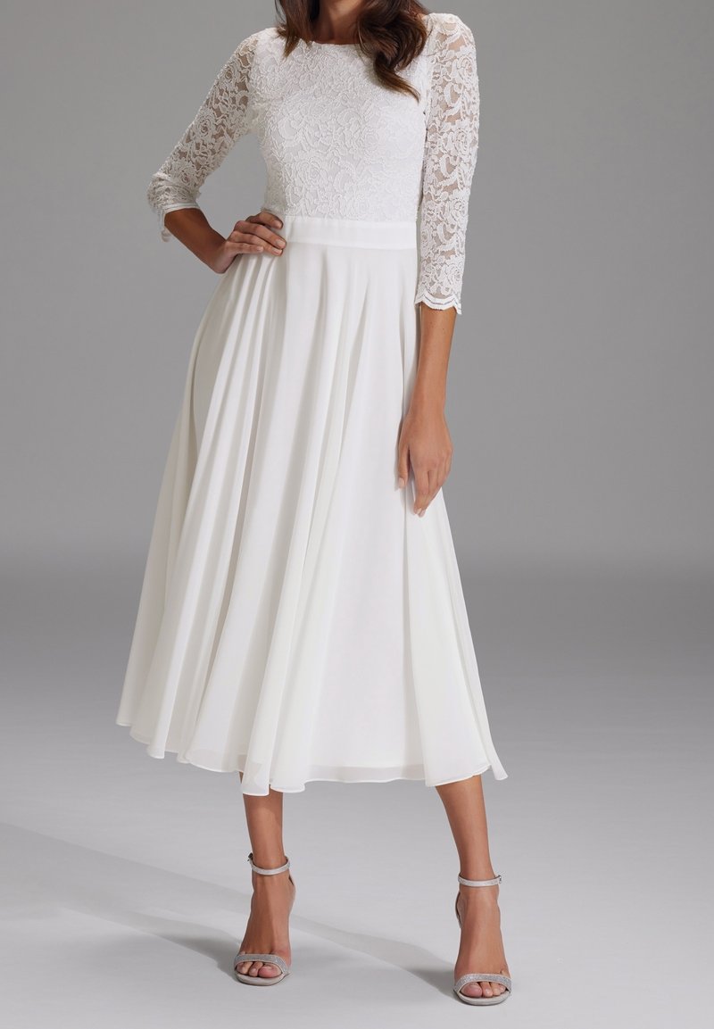 Femme portant une robe midi blanche avec un corsage ajusté en dentelle et des manches trois-quarts, associée à des talons argentés à bride à la cheville, debout.