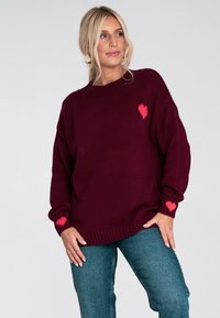 Burgundzkie sweter z dzianiny z okrągłym dekoltem, z małym różowym serduszkiem na piersi i przy mankietach, zestawione z niebieskimi dżinsami.