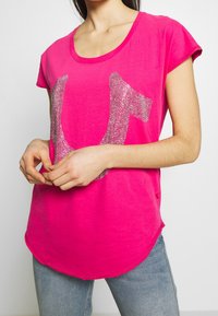 Mujer vistiendo una camiseta de manga corta rosa brillante con un gran diseño de herradura de circonio plateado, combinada con jeans azul claro, con las manos entrelazadas al frente.