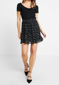 Haut noir à côtes avec un décolleté en V et une jupe plissée noire ornée de pois blancs. La tenue est complétée par des talons noirs à lanières.