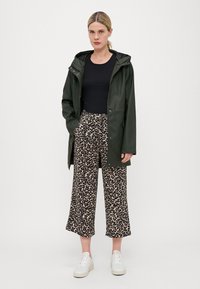 Mørkegrønn vanntett jakke med hette, svart topp og løse beige leopardmønstrede bukser. Hvite sneakers fullfører antrekket.