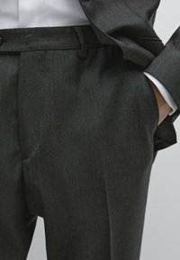 Mann in dunkelgrauen Anzughose mit der Hand in der Tasche, trägt ein weißes Hemd und eine passende Anzugjacke.