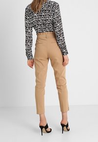 Kvinde iført højtaljede beige skræddersyede bukser, en langærmet top med sort-hvidt mønster og sorte åbne højhælede sko, stående foran en hvid væg.