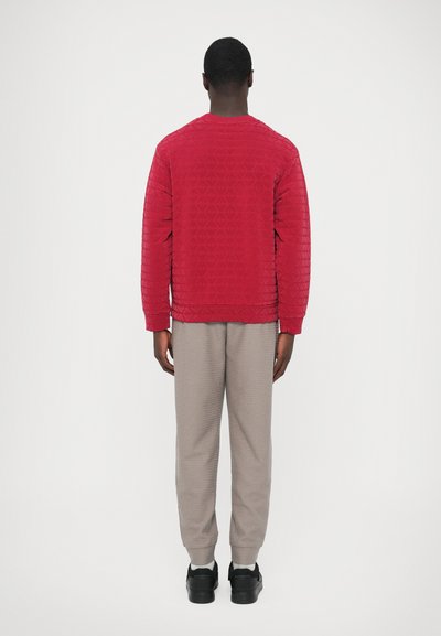 Κόκκινο κουβερταριστό φούτερ με γεωμετρικό μοτίβο, συνδυασμένο με ανοιχτό γκρι υφασμένα παντελόνια και μαύρα παπούτσια, από πίσω όψη.