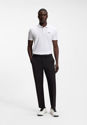 Man draagt een witte polo met korte mouwen en het "BOSS"-logo, zwarte broek en witte sneakers met zwarte veters, staand met handen in de zakken.