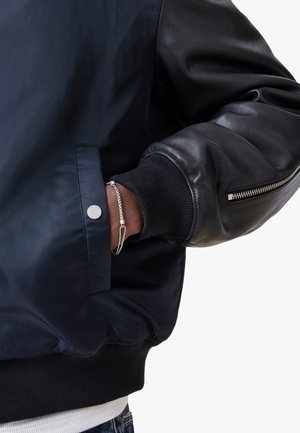 Giacca bomber blu navy con design a maniche in pelle, caratterizzata da polsini a coste, tasca con zip e dettaglio con bottone a pressione metallico.