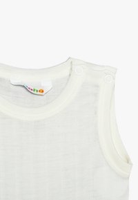 Ljusgräddfärgad ärmlös topp gjord av mjukt, texturerat tyg med två tryckknappar på axeln och ribbat halsringningsdetalj.