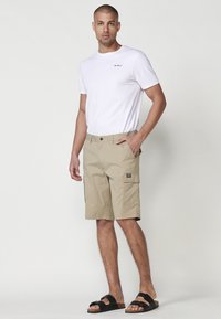 Tricou alb din bumbac cu logo brodat, asociat cu pantaloni scurți bej din material cargo cu buzunare laterale și sandale negre cu talpă din plută.