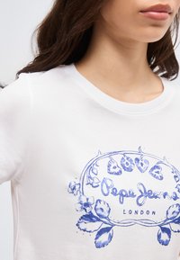 Γυναίκα που φοράει λευκό μπλουζάκι με μπλε ανθικό σχέδιο και το κείμενο "Pepe Jeans London" στο στήθος.