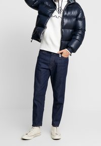 Chaqueta acolchada de color navy con acabado brillante, sudadera blanca con capucha y logo, pantalones de mezclilla oscura y zapatillas altas beige.