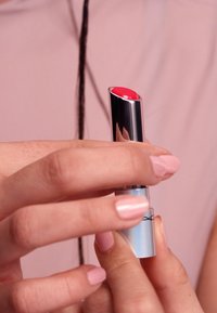 Lipstick in a silver tube with a glossy finish, featuring a round red tip. Hand holding it with light pink nails against a plain background.