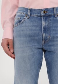 Lyseblå denimjeans med klassisk design, som har knappelukking, fem lommer og synlige sømdetaljer.