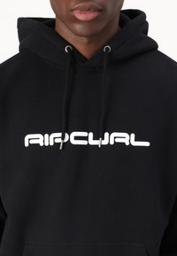 Hanorac negru cu un mare logo alb "Ripcurl". Dispune de șnururi cu capete argintii și o textură moale. Croială normală cu un buzunar frontal.
