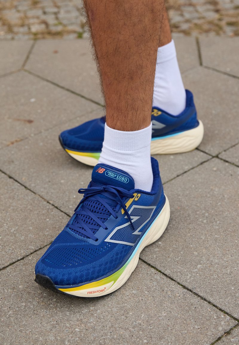 Chaussures de sport bleues avec tige en mesh, présentant des accents réfléchissants, des lacets blancs et une semelle rembourrée de plusieurs couleurs. Portées avec des chaussettes blanches.