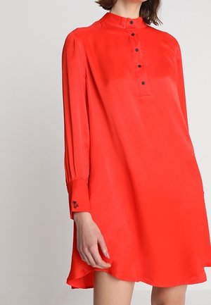 Frau trägt ein locker sitzendes rotes Langarmkleid mit Mandarin-Kragen und schwarzen Knöpfen auf der vorderen Blende und den Manschetten.