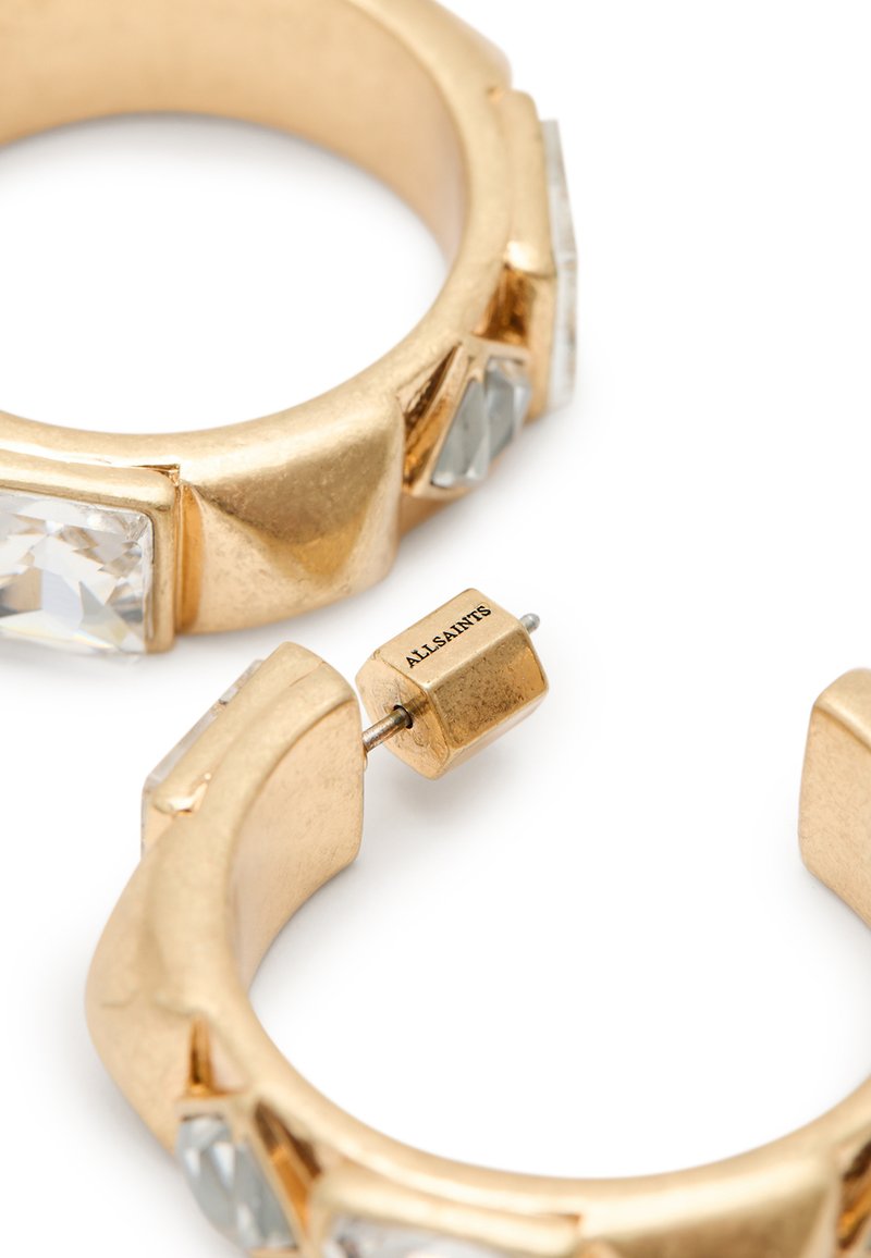 Goldene Creolenohrringe mit klaren, facettierten Steinen und glatten Oberflächen. Hervorzuhebende Details sind die eckigen Formen und ein markiertes Schließsystem.