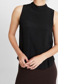 Czarny top bez rękawów z wysokim dekoltem, wykonany z gładkiego materiału. Lekko luźny krój z zaokrąglonym dołem, zestawiony z teksturowaną brązową spódnicą.